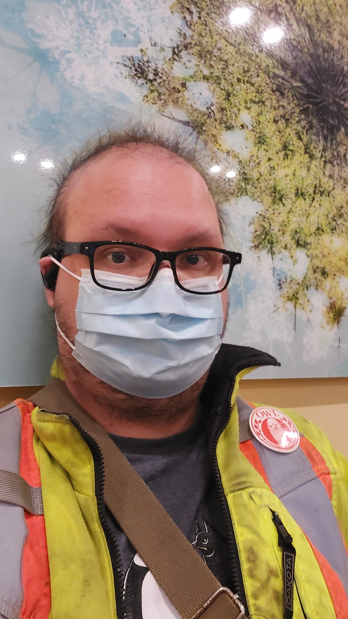 A man with thin hair wearing a high visibility coat and surgical mask sits in the waiting room of a hospital